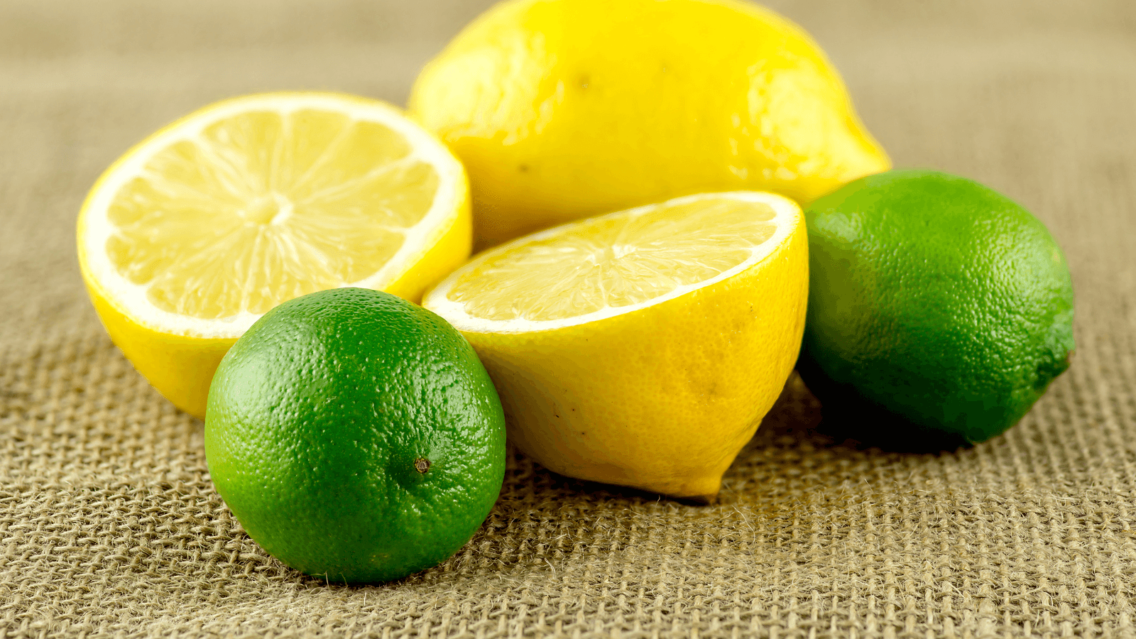Fresh whole and halved lemons and limes displayed on a burlap surface.