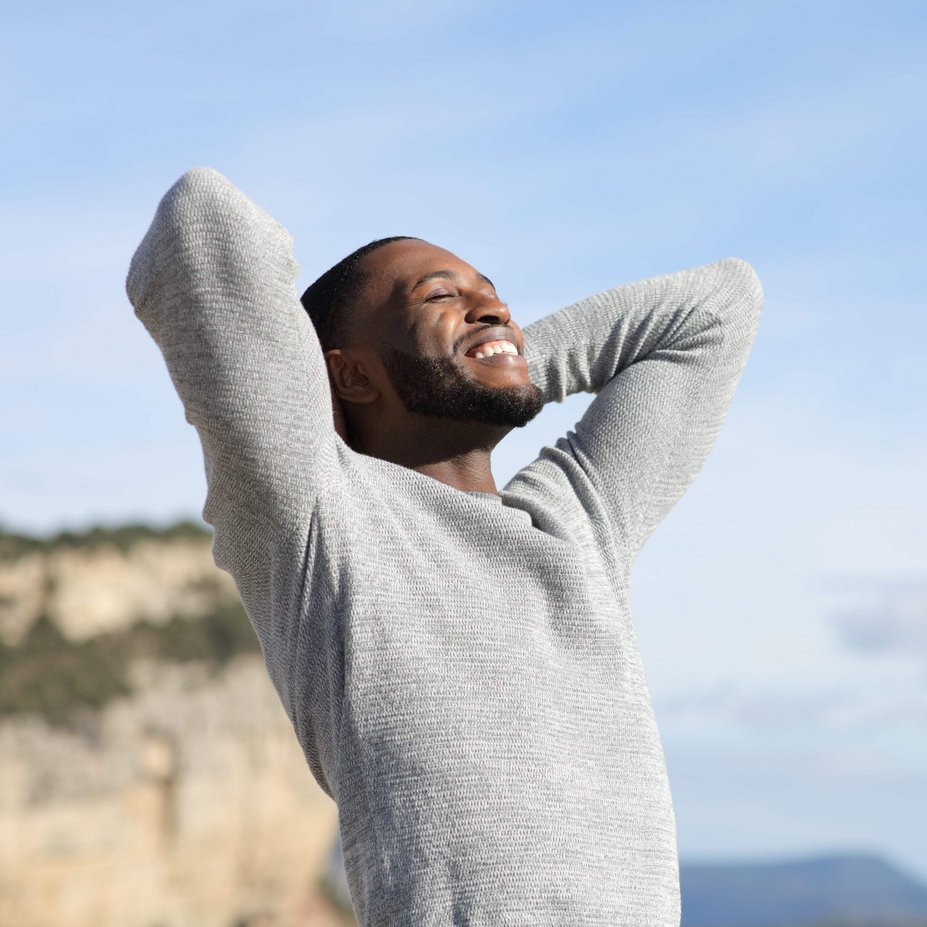 Smiling man standing outdoors with hands behind head, enjoying fresh air and nature.
