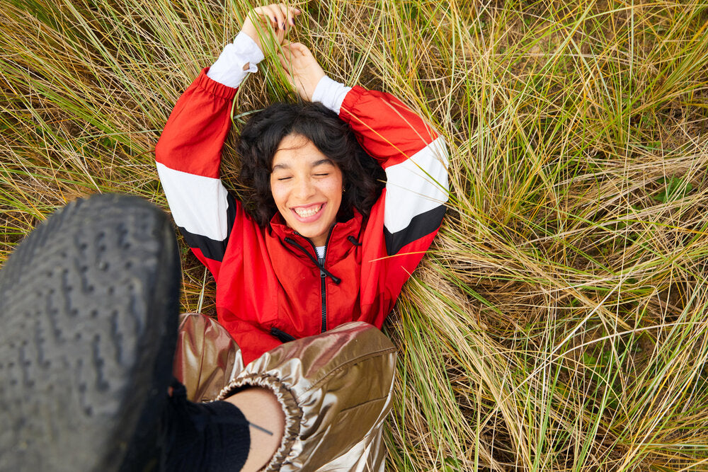 Smiling person lying in tall grass wearing a red jacket and gold pants, holding hands up playfully.