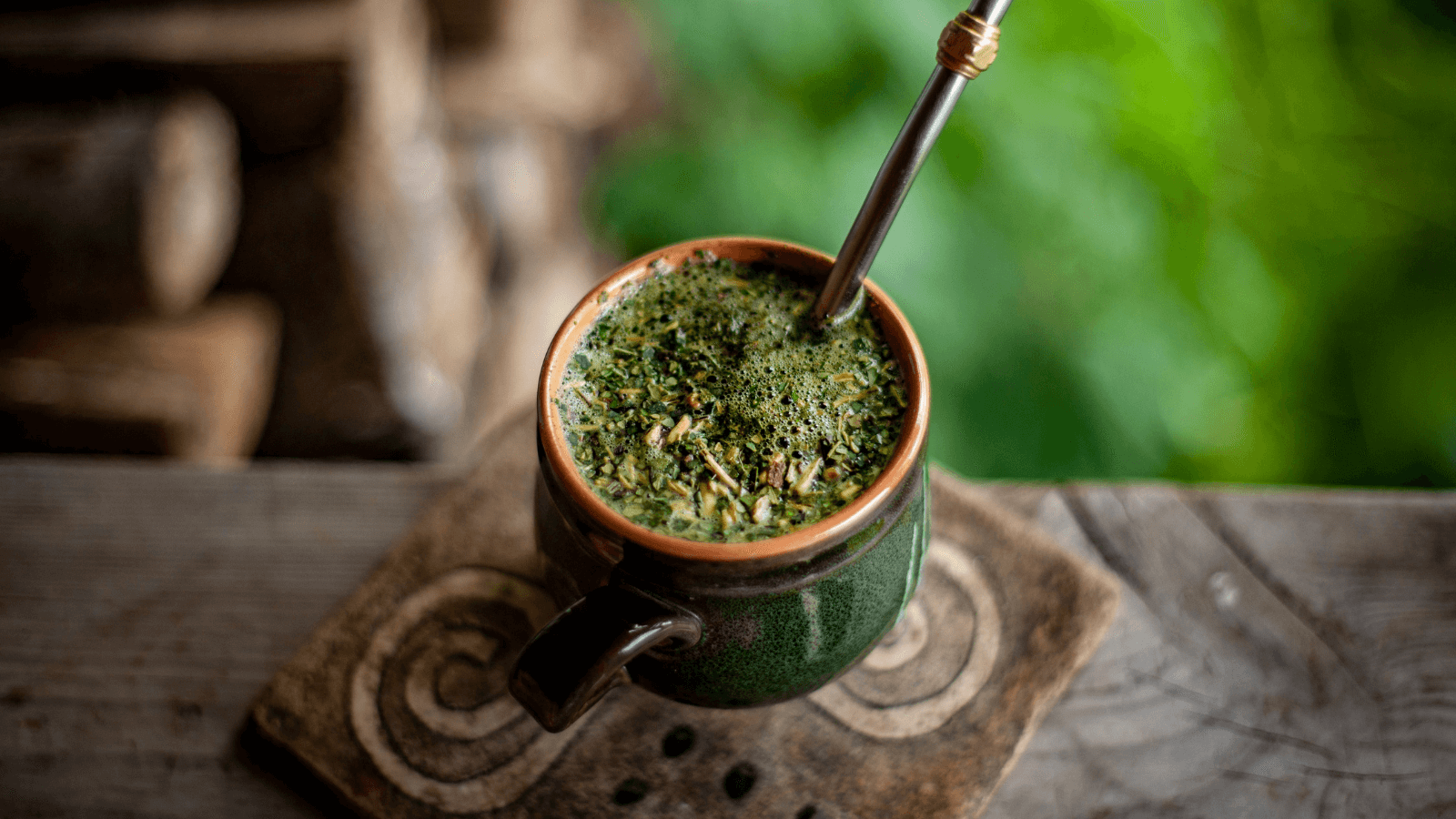 Ceramic mug of yerba mate with a metal straw, sitting on a wooden surface with blurred greenery in the background.